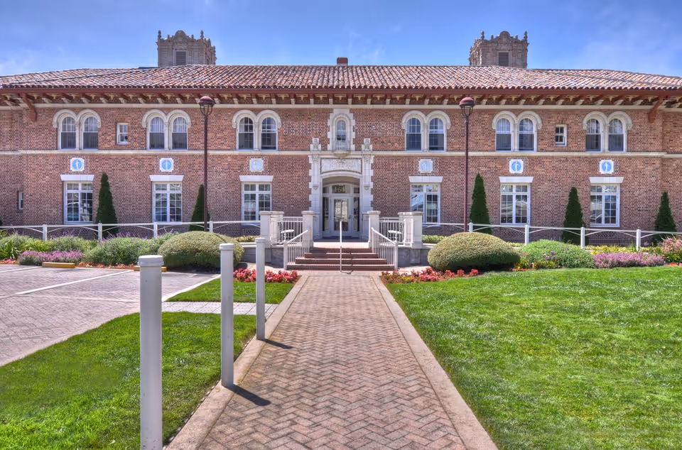 Front exterior view of a large brick building with a tiled roof, multiple windows, and decorative architectural details. A paved walkway leads to the main entrance, flanked by manicured bushes, flower beds, and green lawns under a clear blue sky.