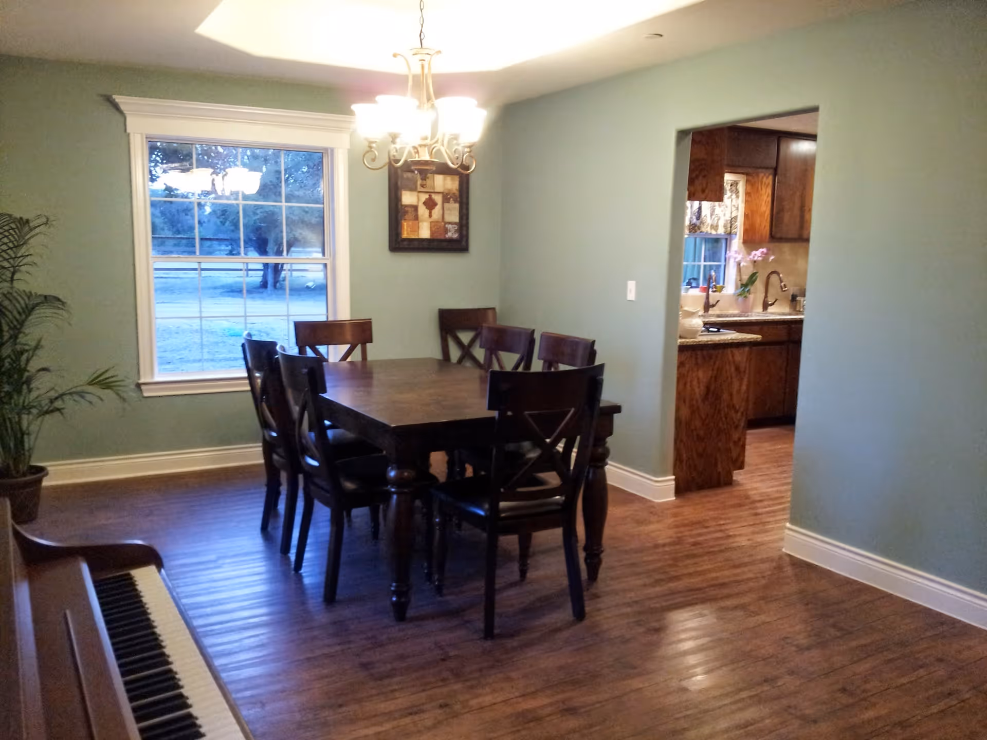 A dining room with a wooden dining table and six matching chairs. A chandelier hangs above the table. There is a large window with a view of trees outside, a framed artwork on the wall, and a potted plant in the corner. To the right, there is an open doorway leading to a kitchen with wooden cabinets and a countertop with a sink.