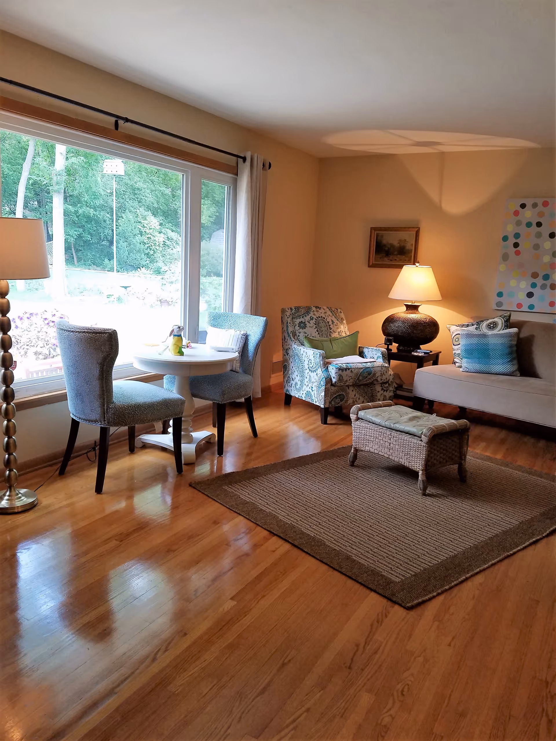 Sunlit living room with hardwood floors, a large window, a small round table with chairs, an armchair and sofa with a side table and lamp, and a woven ottoman on a rug.