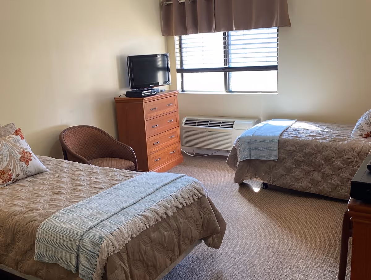 Twin-bed bedroom with two beds, a dresser topped by a TV, a chair, and a window with blinds.