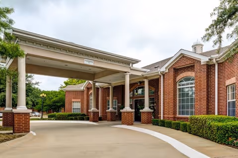 Front entrance of a red-brick senior living building with a covered porte-cochere, large arched windows, and trimmed hedges.