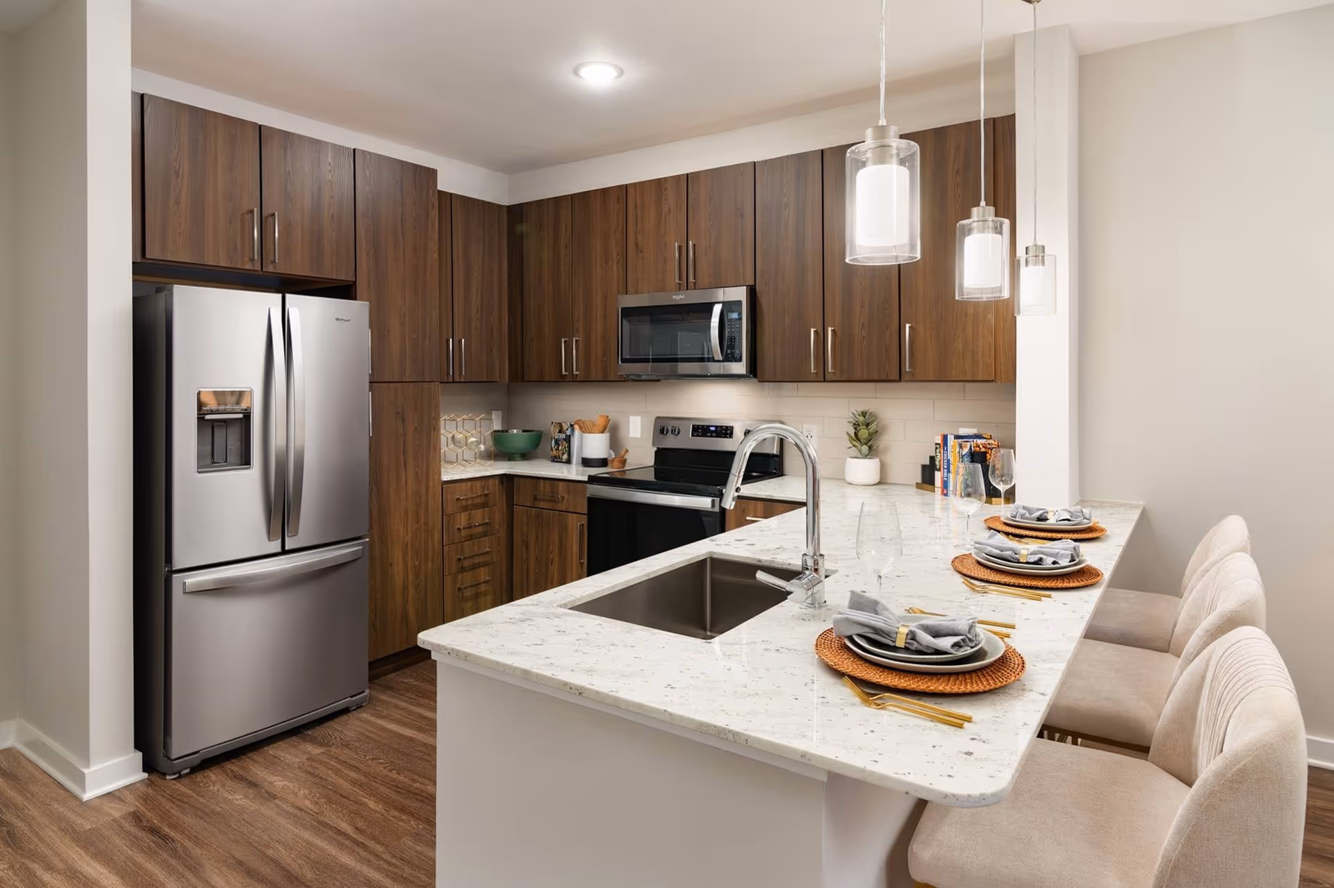 Modern kitchen featuring a marble island with sink and three set bar stools, stainless steel refrigerator and appliances, wood cabinets, and pendant lighting.