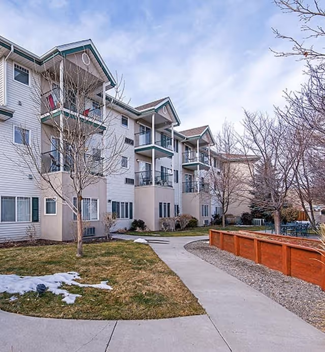 Exterior view of a multi-story residential building with balconies, surrounded by leafless trees and a pathway leading through a grassy area with patches of snow.