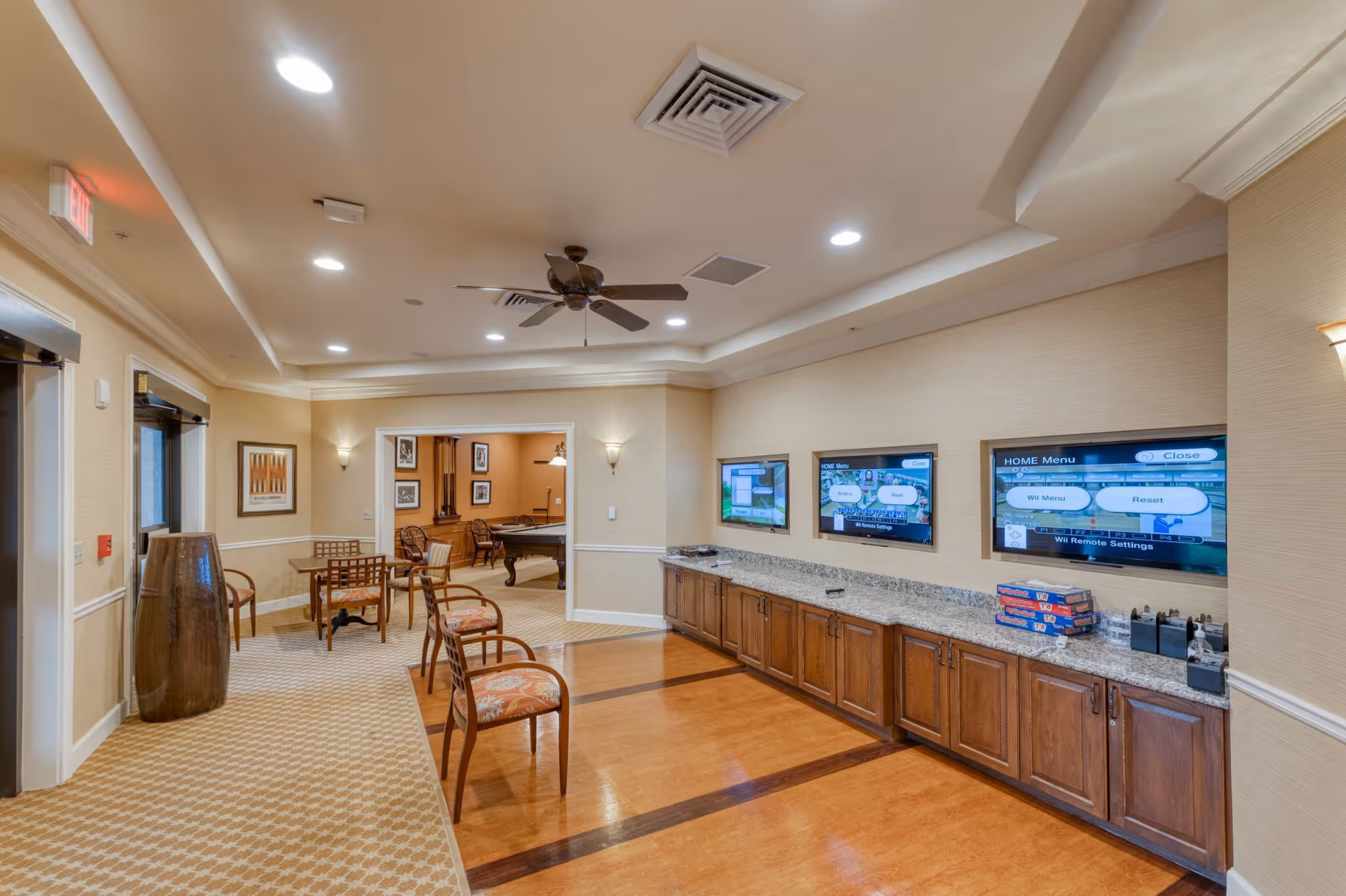 A spacious common area in a senior living facility featuring a seating area with chairs and a table, a long granite countertop with wooden cabinets underneath, and three wall-mounted screens displaying a Wii game menu. In the background, there is a room with a pool table and framed pictures on the walls. The ceiling has recessed lighting and a ceiling fan.