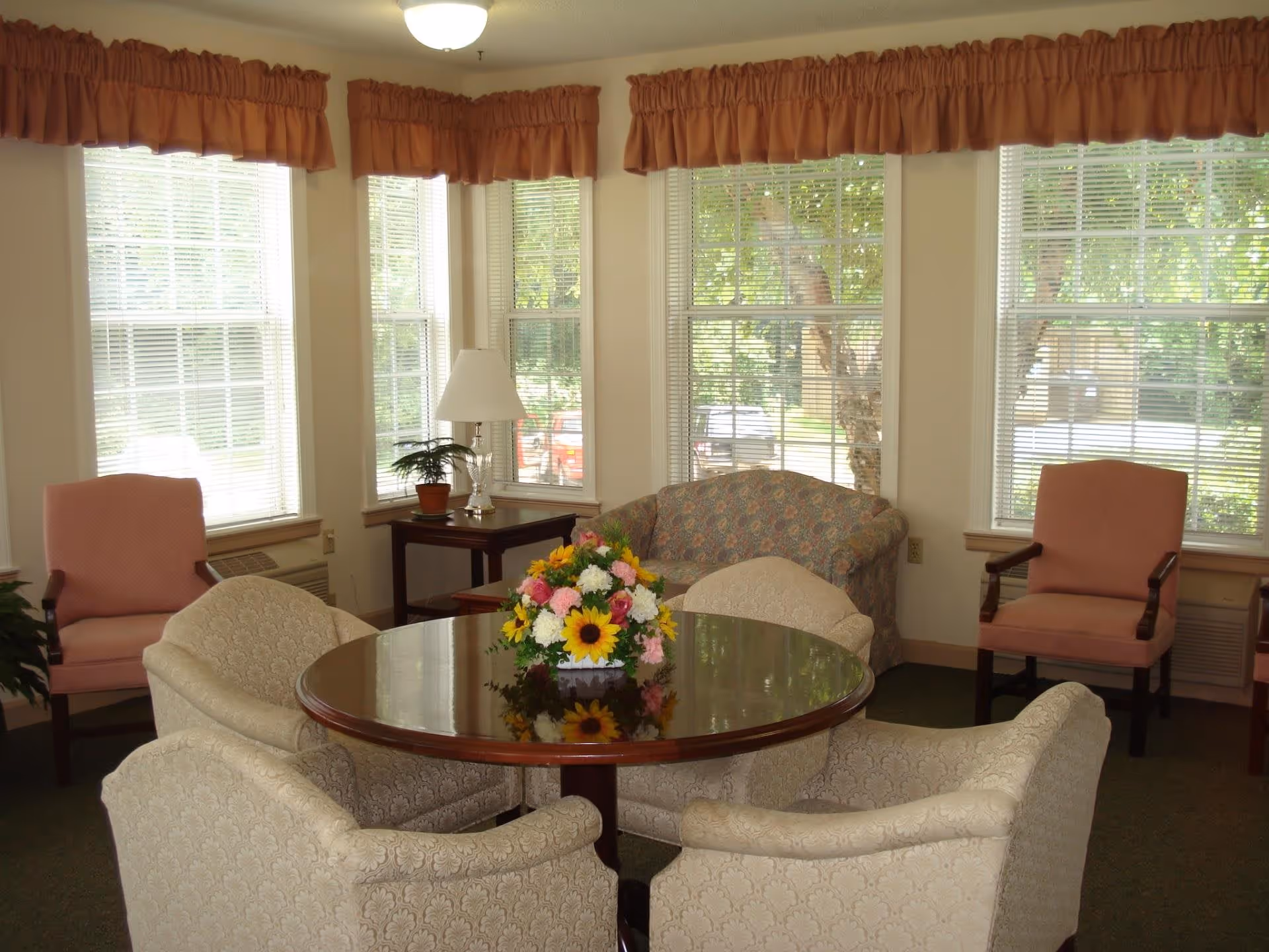 A cozy sitting area with a round wooden table surrounded by four upholstered armchairs. On the table is a colorful floral arrangement. Behind the table, there is a floral-patterned loveseat and two pink upholstered chairs near large windows with white blinds and orange valances. A small side table with a lamp and a potted plant is also visible.
