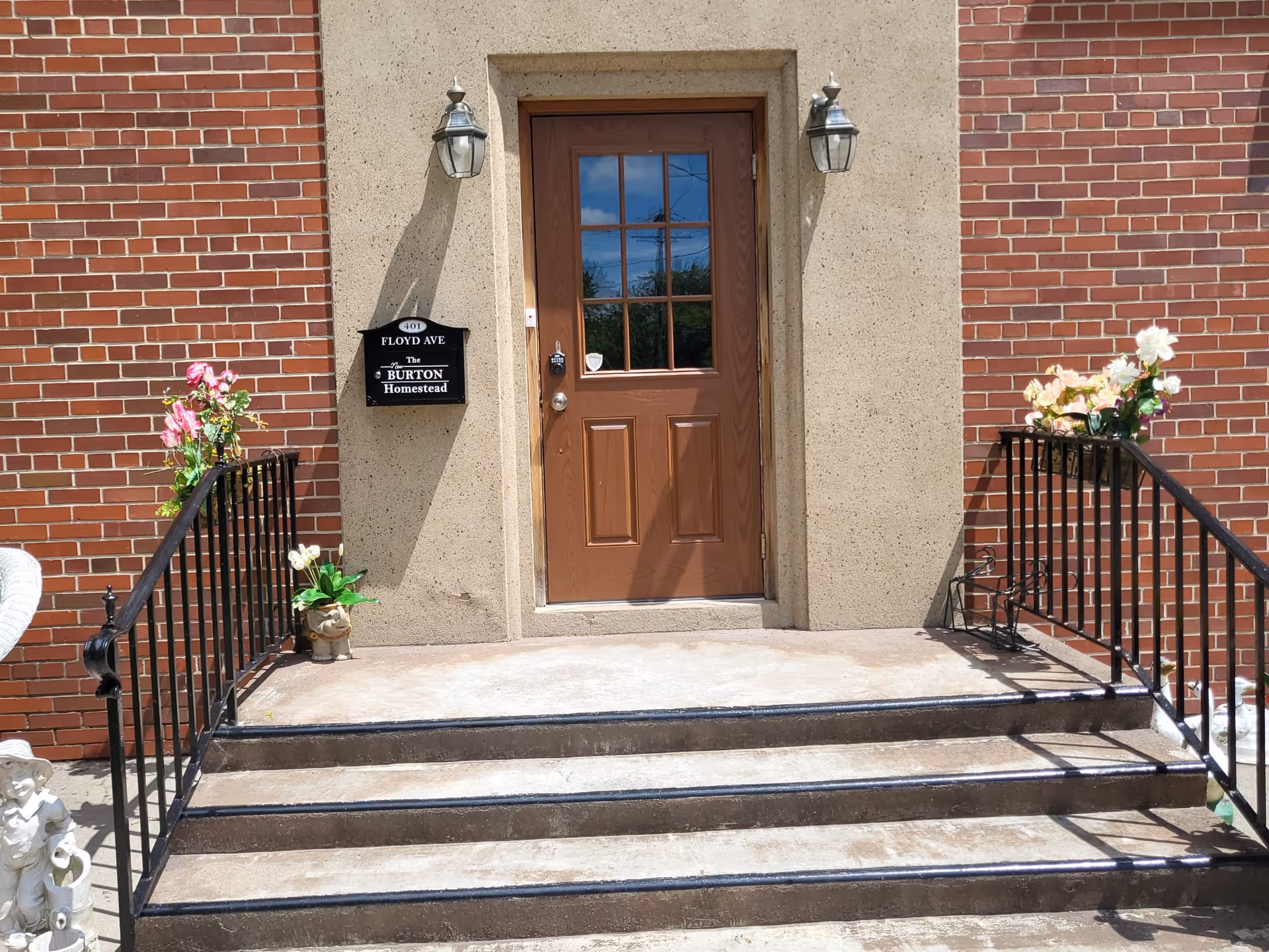 Front entrance of a brick building with a brown door, concrete steps with black metal railings, potted flowers, and a sign reading 'Burton Homestead'.