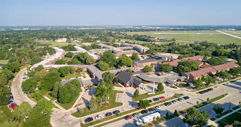 Aerial view of Luther Manor senior living facility showing multiple buildings surrounded by trees and greenery, with cars parked along the roads and a large open field in the background.