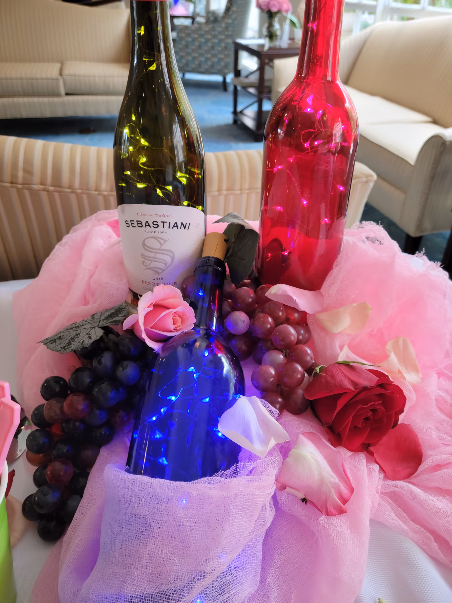 Table centerpiece with blue, pink, and green decorative bottles, string lights, faux grapes and roses arranged on pink fabric in a seating area.