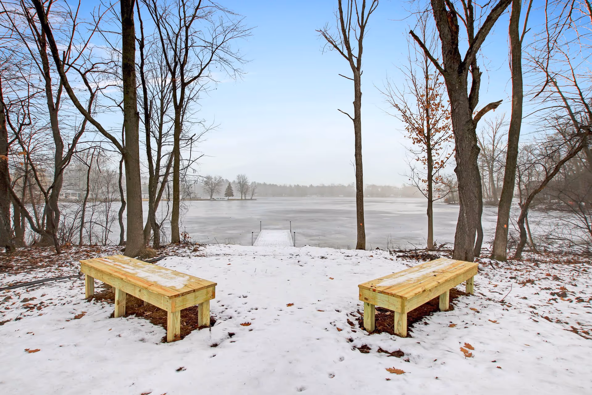 Snow-covered ground with two wooden benches facing a frozen lake surrounded by leafless trees under a clear sky.