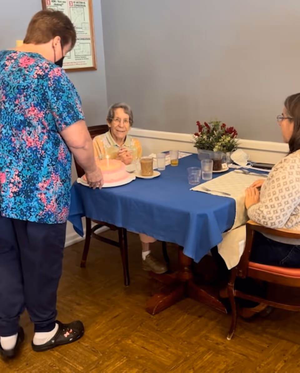 An elderly woman sitting at a table covered with a blue tablecloth, smiling and looking at a woman standing nearby who is holding a pink birthday cake with lit candles. Another woman is seated at the table, facing the elderly woman. The table has several cups, a flower arrangement, and a placemat.