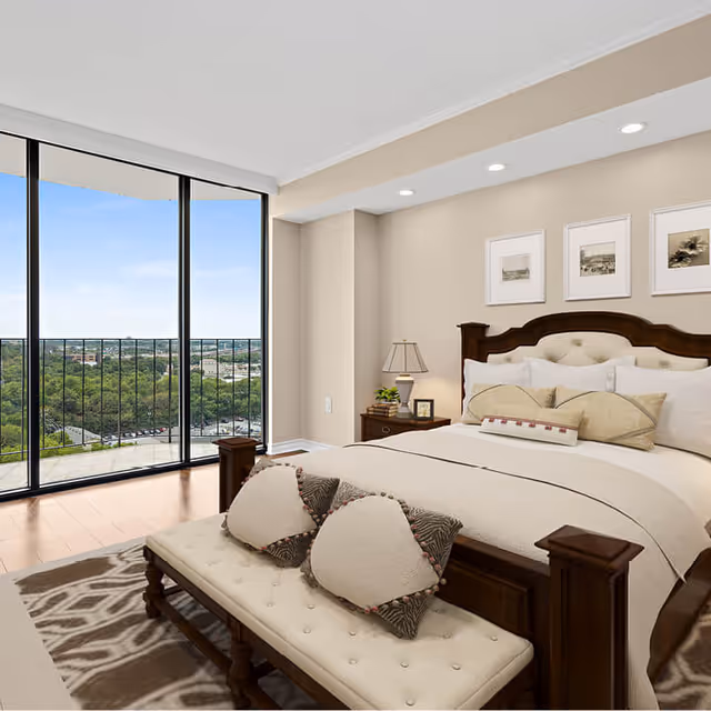 A well-lit bedroom with a large bed featuring multiple pillows and a wooden headboard. There is a cushioned bench with decorative pillows at the foot of the bed. A nightstand with a lamp, small plant, and framed photo is beside the bed. The room has light-colored walls with three framed pictures above the headboard. Floor-to-ceiling windows open to a balcony with a view of trees and a distant cityscape under a clear blue sky.