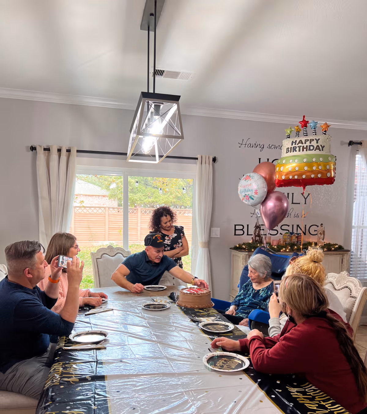 A family gathered around a dining table as a man presents a birthday cake to an elderly woman with balloons and decorations in the background.