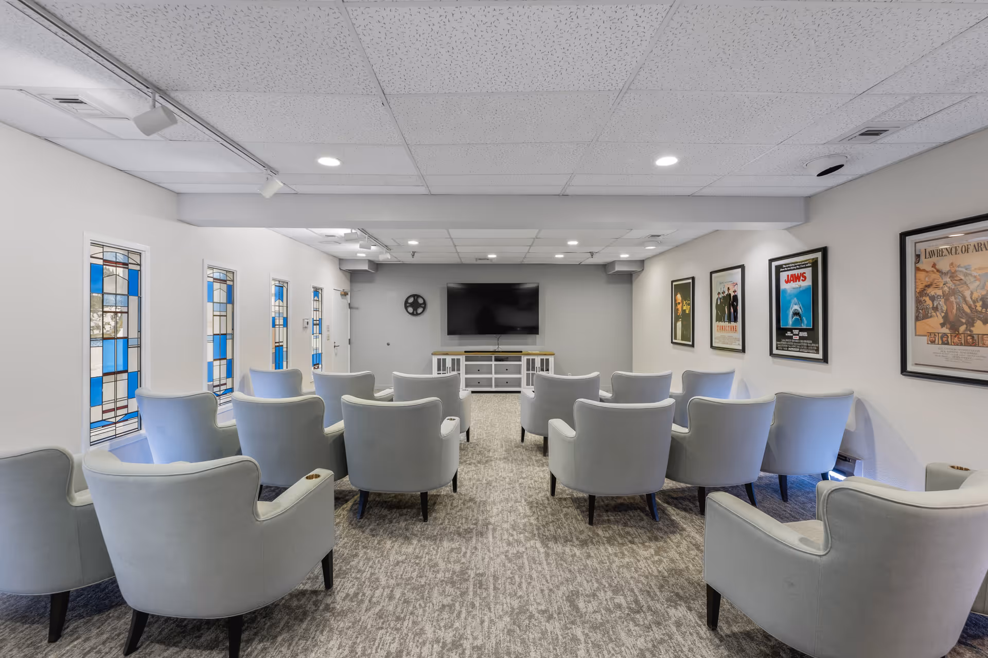 A cozy media room with multiple light gray armchairs arranged in rows facing a large flat-screen TV mounted on the wall. The room has a carpeted floor, white walls adorned with framed movie posters, and decorative stained glass windows on one side.