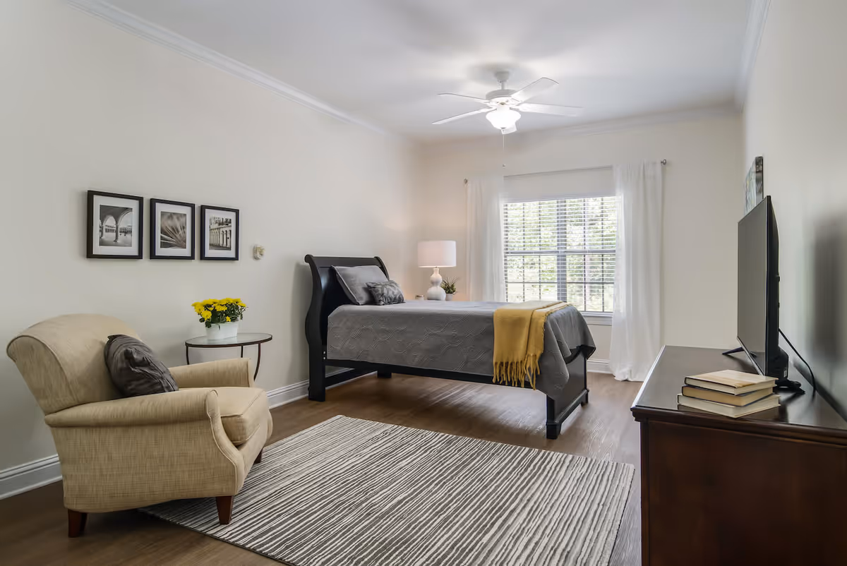 A bright, simply furnished bedroom with a bed dressed in gray linens and a yellow throw, an armchair, TV on a dresser, and a window with white curtains.