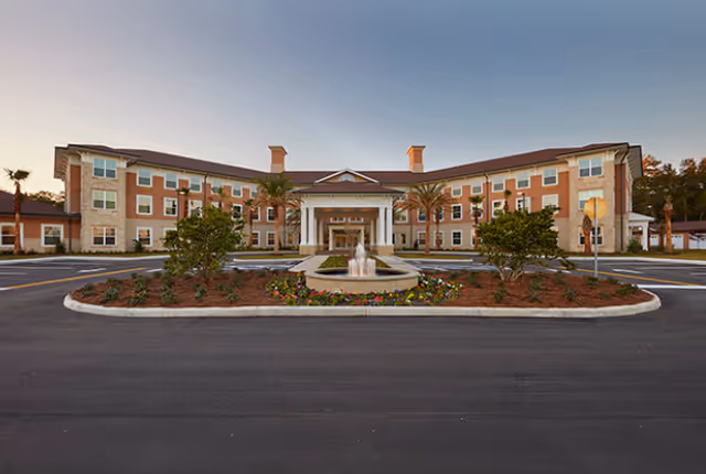 Front exterior view of Canterfield of Ocala, a three-story building with a central entrance featuring columns, a fountain in front, palm trees, and landscaped flower beds.