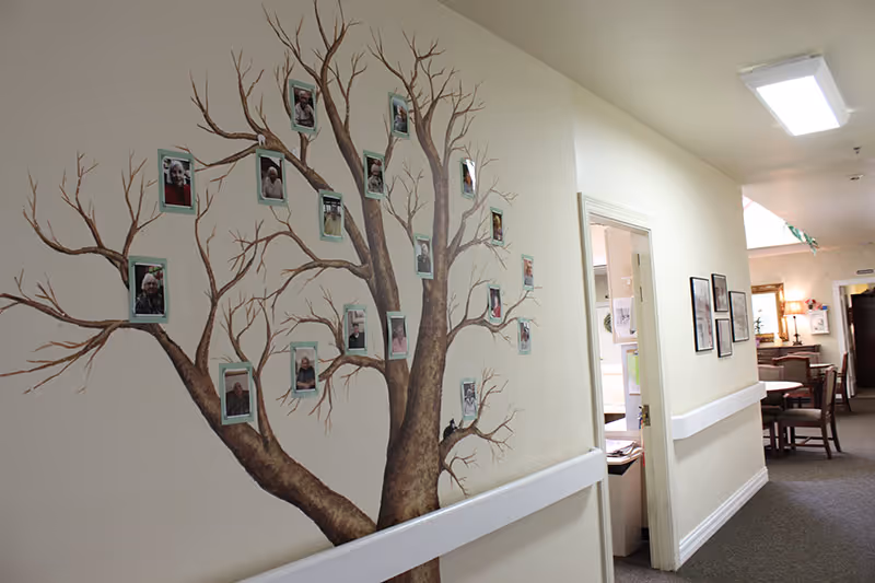 Interior hallway of a senior living facility with a large painted tree on the wall displaying framed photos of residents. The hallway has beige walls, a white chair rail, and carpeted floor. There are open doorways and framed pictures on the opposite wall, with tables and chairs visible further down the hall.