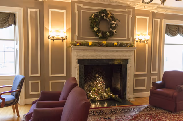 A cozy living room with a decorated fireplace featuring a wreath and garland. There are two maroon armchairs and a maroon loveseat arranged around a patterned rug. The walls have decorative molding and two wall sconces with lights. Two windows with valances allow natural light into the room.