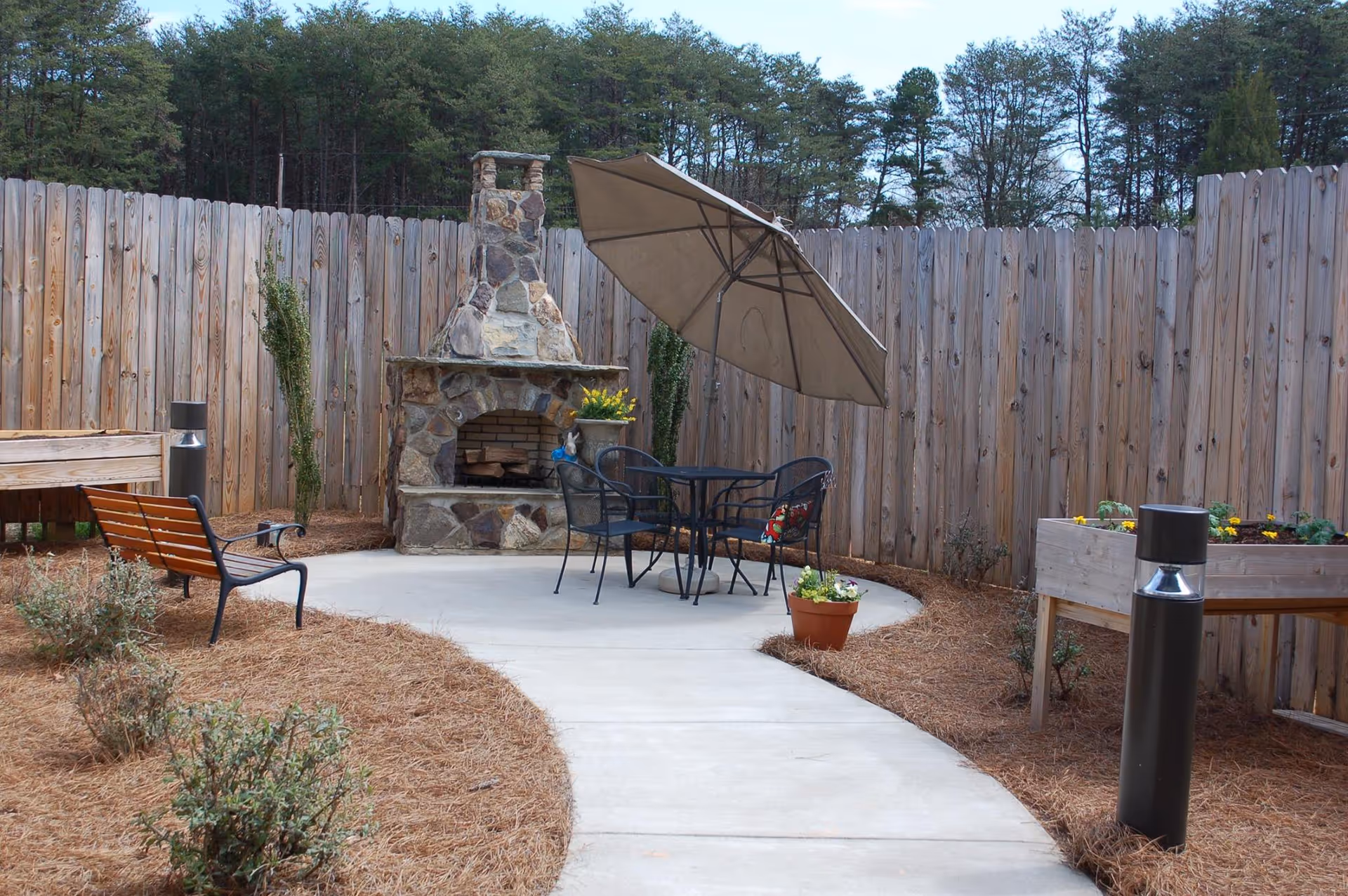 Outdoor patio area with a stone fireplace, a round metal table with four chairs, and a large beige umbrella. The space is enclosed by a tall wooden fence, with some plants and shrubs along the edges and a concrete pathway leading to the seating area.