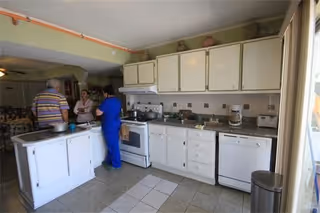 Kitchen interior with white cabinets, a stove and dishwasher, an island, and three people talking near the counter.