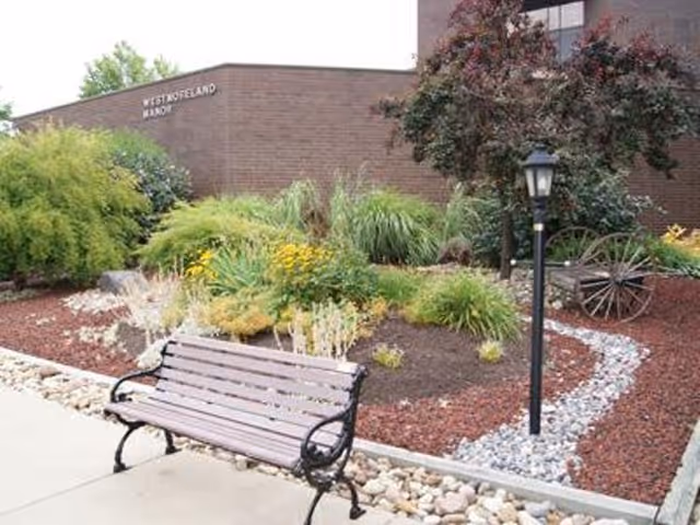 Landscaped front garden with a bench, lamp post, and decorative wagon wheel in front of a brick building labeled Westmoreland Manor.