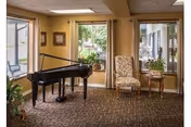 A bright communal living room with a black grand piano, upholstered chairs, a side table with a plant, and large windows looking outside.