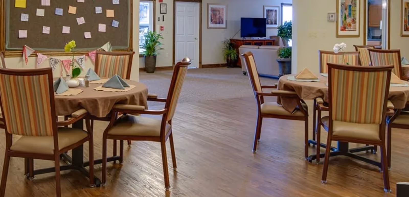 A senior living facility dining area with round tables covered in brown tablecloths, each set with folded napkins and small decorative items. The chairs have striped upholstery. In the background, there is a bulletin board with notes pinned on it, a TV on a stand, some potted plants, and framed artwork on the walls.