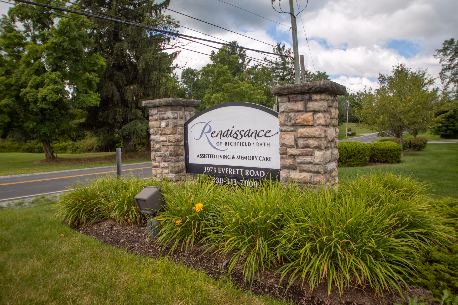 Stone sign for Renaissance of Richfield/Bath assisted living and memory care facility surrounded by green plants and grass, with trees and a road in the background under a partly cloudy sky.