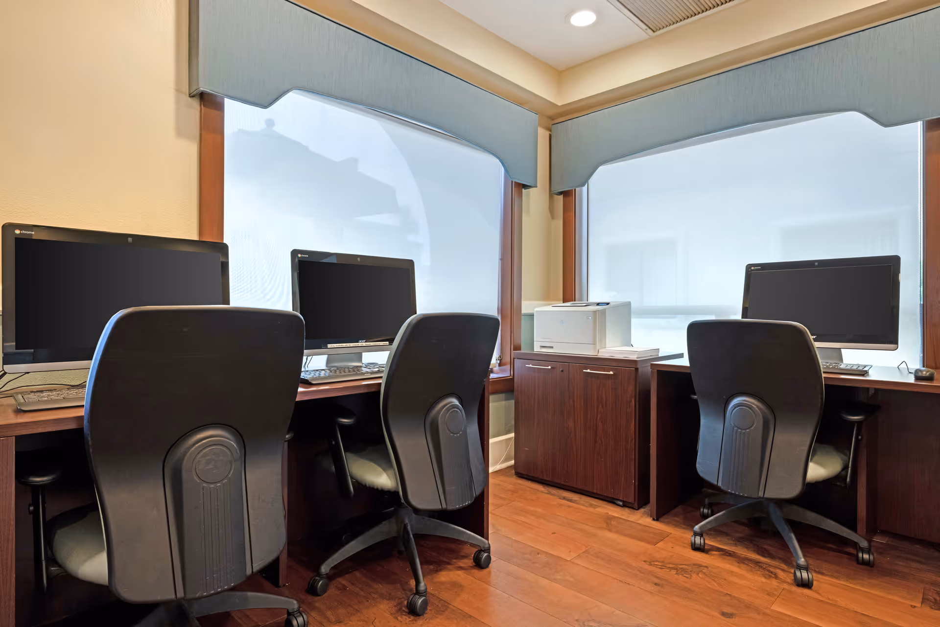 A small office or computer room with three black office chairs and three desktop computers on wooden desks. There is a wooden cabinet with a printer on top placed between two windows with light blue valances. The floor is wooden.