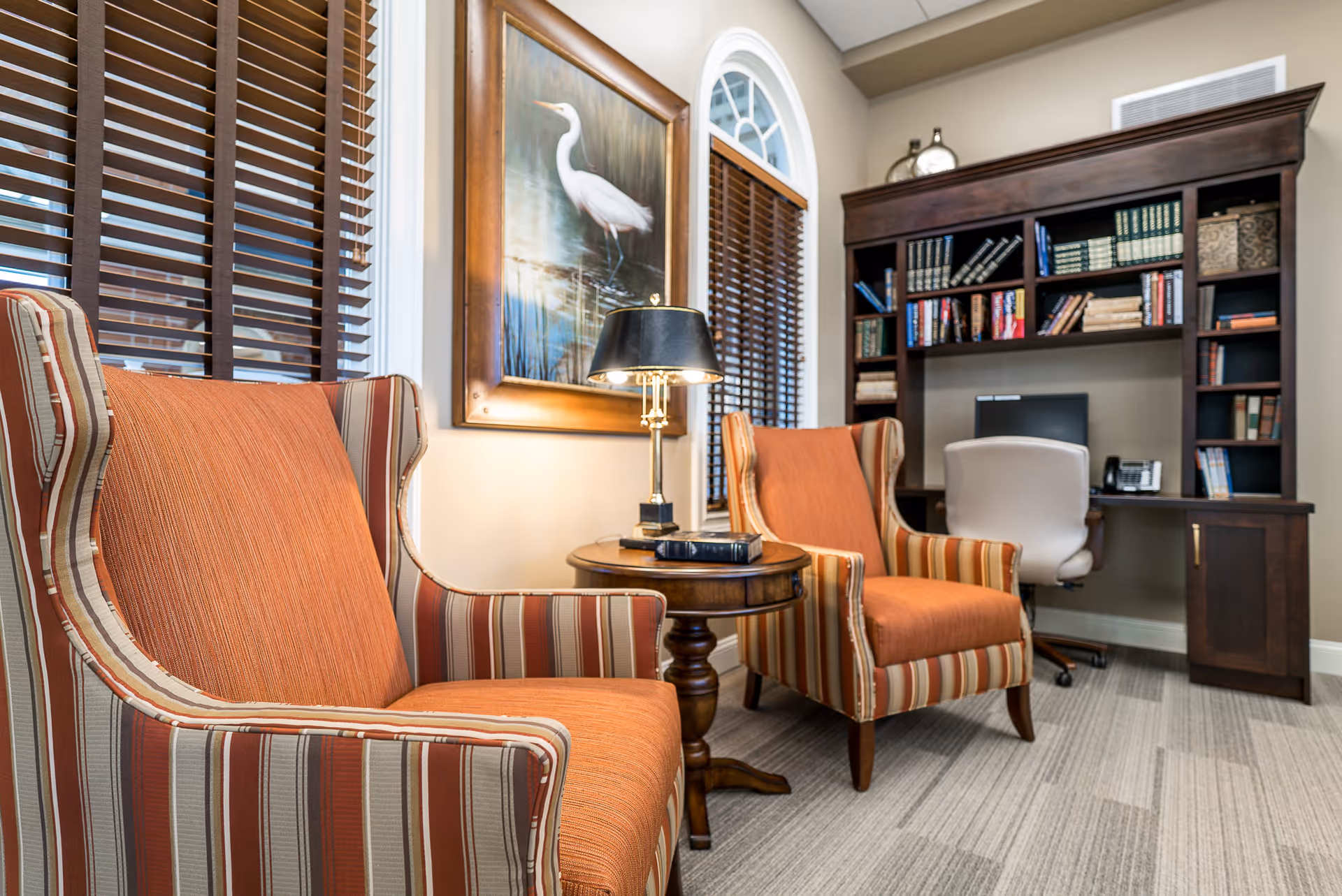A cozy interior room with two striped armchairs featuring orange cushions, a round wooden side table with a black lamp and a book, a large framed painting of a white heron on the wall, and a dark wooden bookshelf with a computer and office chair.