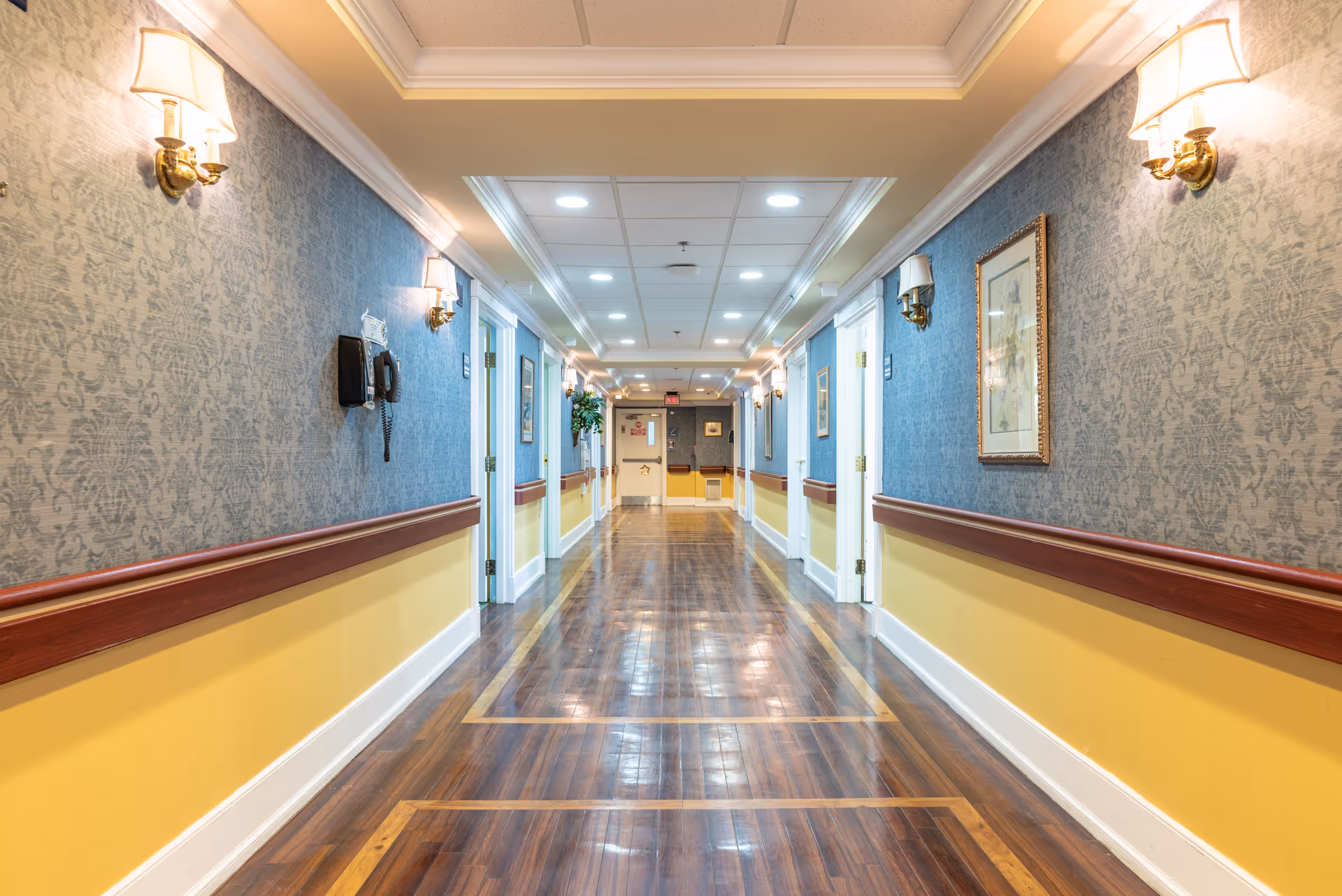 A well-lit hallway in a senior living facility with polished wooden floors, blue patterned wallpaper above a yellow wainscoting, wall-mounted lamps, framed artwork, and several doors along the corridor.