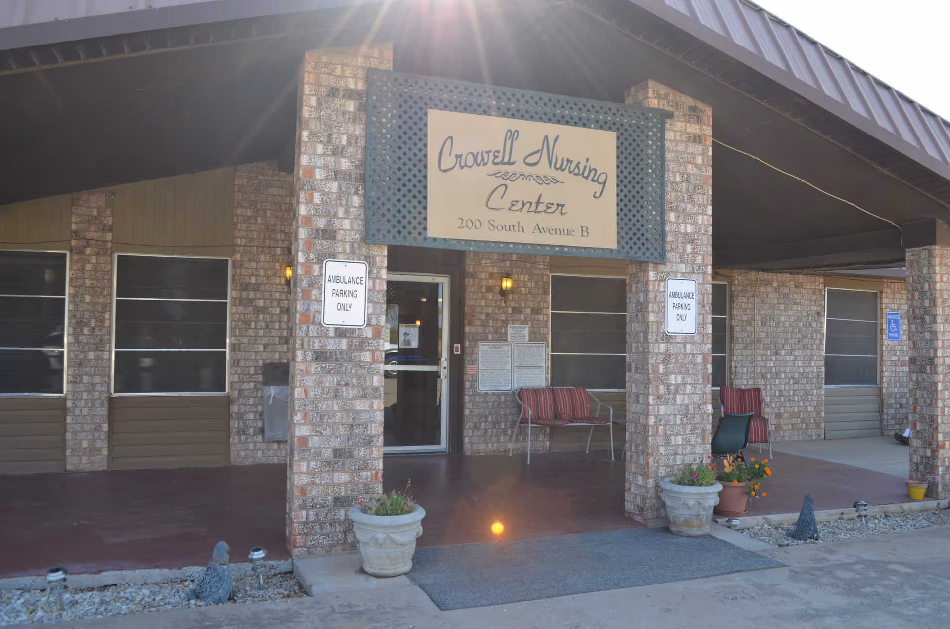 Entrance of Crowell Nursing Center with a covered porch supported by brick pillars. There are two benches with red cushions, potted plants, and signs indicating ambulance parking only. The building has a brick exterior and several windows with screens.