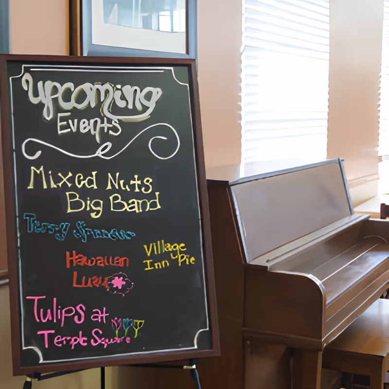 A chalkboard sign listing upcoming events including Mixed Nuts Big Band, Terry Spence, Hawaiian Luau, Village Inn Pie, and Tulips at Temple Square, placed next to a wooden piano in a room with large windows covered by white blinds.