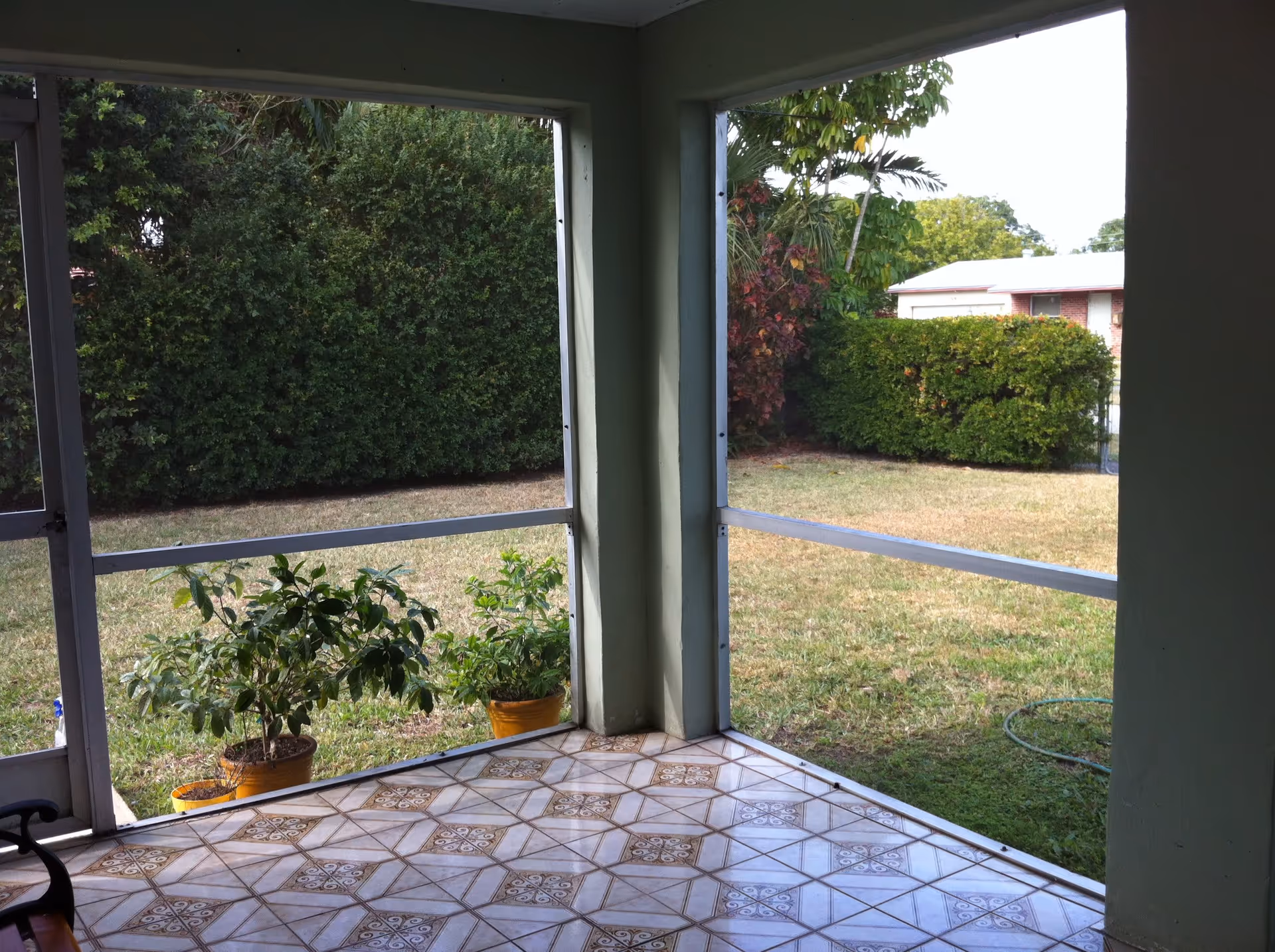 View from a tiled porch or patio area looking out onto a grassy yard with bushes and trees. There are two potted plants on the edge of the porch, and a garden hose is visible on the grass. The porch is screened in with metal framing.