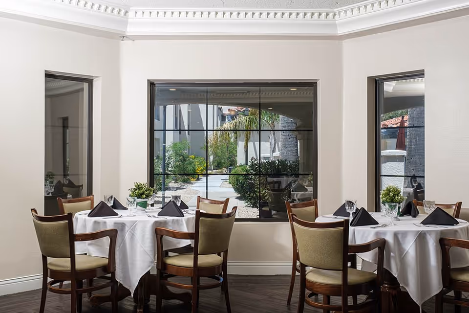 Dining room with round tables covered in white tablecloths, wooden chairs, folded black napkins, and large windows looking out to a courtyard.