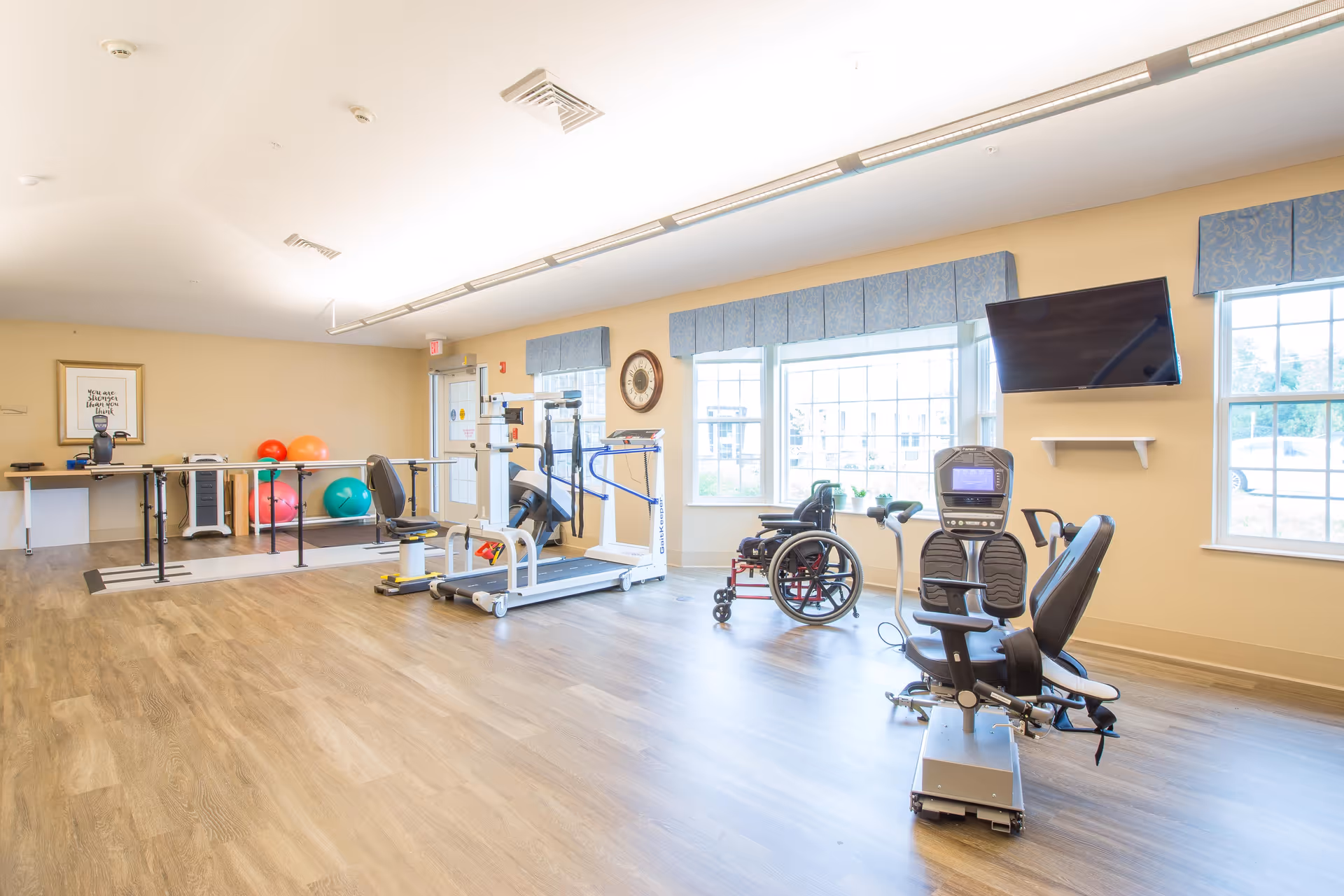 A bright rehabilitation room with exercise equipment including a treadmill, stationary bike, parallel bars, and colorful exercise balls. There is a wheelchair near the windows, a wall-mounted TV, and large windows letting in natural light.