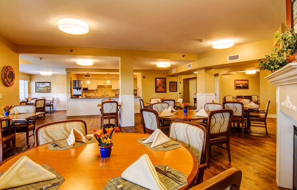 A warm, inviting dining room in a senior living community with multiple round wooden tables set with placemats, folded napkins, and silverware. Each table has a small blue pot with orange flowers. The room features wooden flooring, soft yellow walls, ceiling lights, framed artwork, and a fireplace with a plant on the mantel. In the background, there is a kitchen area with white cabinetry and pendant lights.