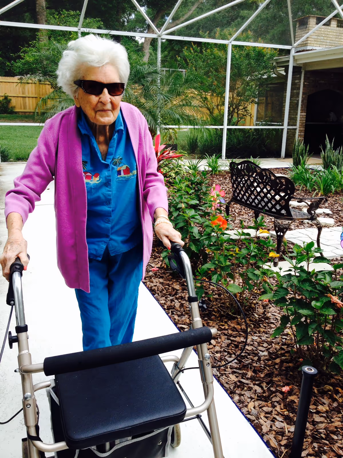 An elderly woman wearing sunglasses, a blue shirt, and a purple cardigan is walking outdoors with a walker on a paved pathway. She is surrounded by a garden with green plants and flowers, and there is a decorative metal bench nearby. The area is enclosed with a screened structure and a wooden fence in the background.