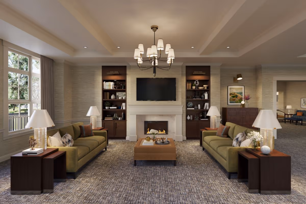 A spacious and well-lit living room with two olive green sofas facing each other, each accompanied by a wooden side table with a lamp and decorative items. In the center is a brown ottoman with books and decor. The room features a fireplace with a mounted flat-screen TV above it, flanked by built-in wooden shelves filled with books and decorative pieces. Large windows on the left allow natural light to enter, and a chandelier hangs from the ceiling. The room has a neutral color palette with beige walls and carpeted flooring.