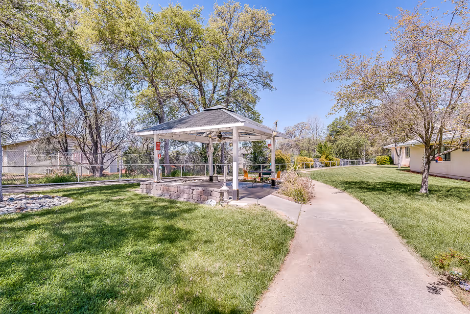 Outdoor area at Meadowood Nursing Center featuring a paved walkway leading to a white gazebo with chairs and tables underneath. The surrounding area has green grass, trees with some leaves, and a clear blue sky.