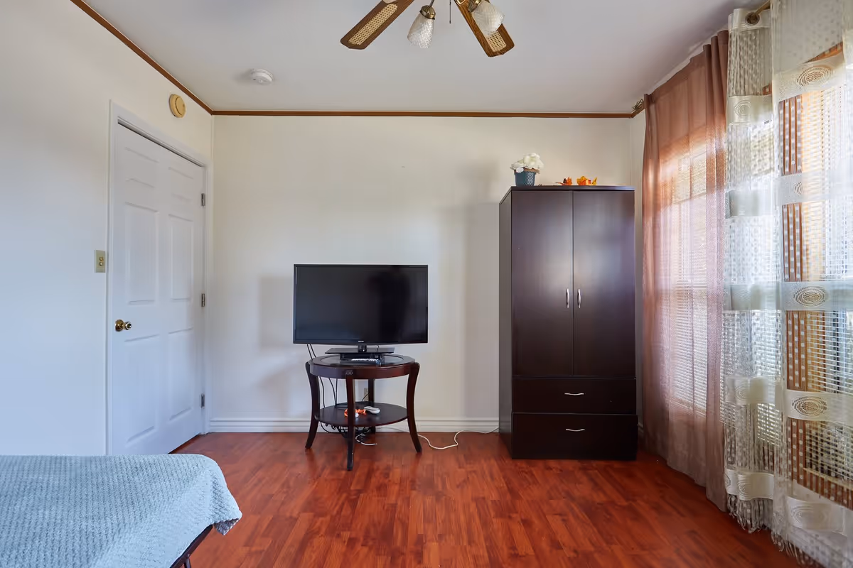 A simple senior living facility room with a wooden floor, a small round table holding a flat-screen TV, a dark wooden wardrobe with two drawers, and a window with sheer and brown curtains allowing natural light to enter. A portion of a bed with a light blue blanket is visible on the left side.