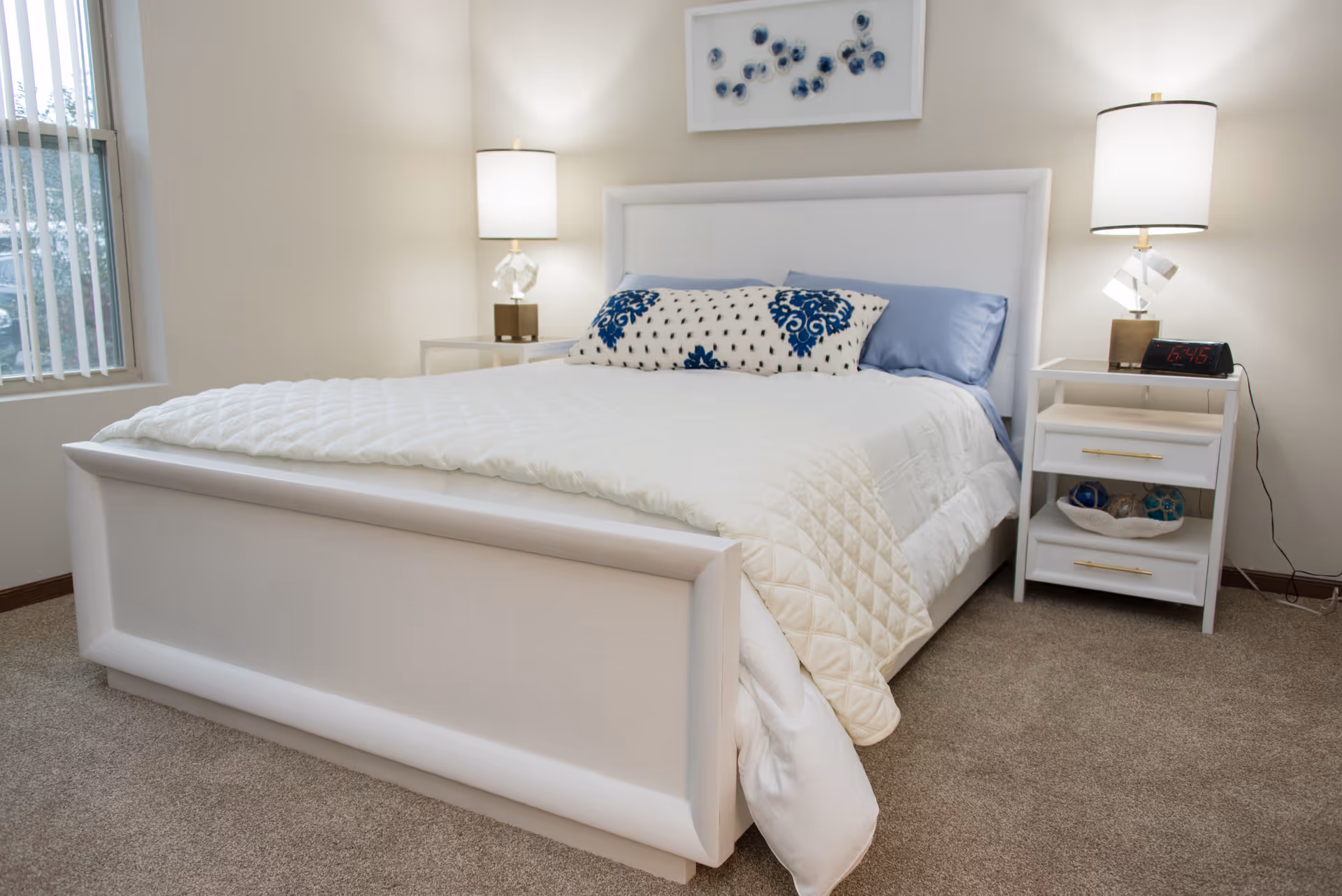 A bright, neatly made bedroom featuring a white upholstered bed flanked by two nightstands with lamps and a window.