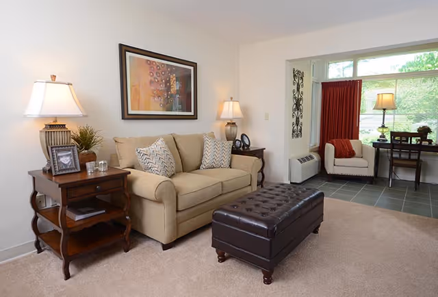 A cozy living room with a beige sofa adorned with patterned cushions, flanked by two wooden side tables each holding a lamp. A framed abstract painting hangs above the sofa. In front of the sofa is a dark brown tufted leather ottoman. In the background, there is a small sitting area with a white armchair, a desk with a lamp, and a large window with red curtains partially drawn.