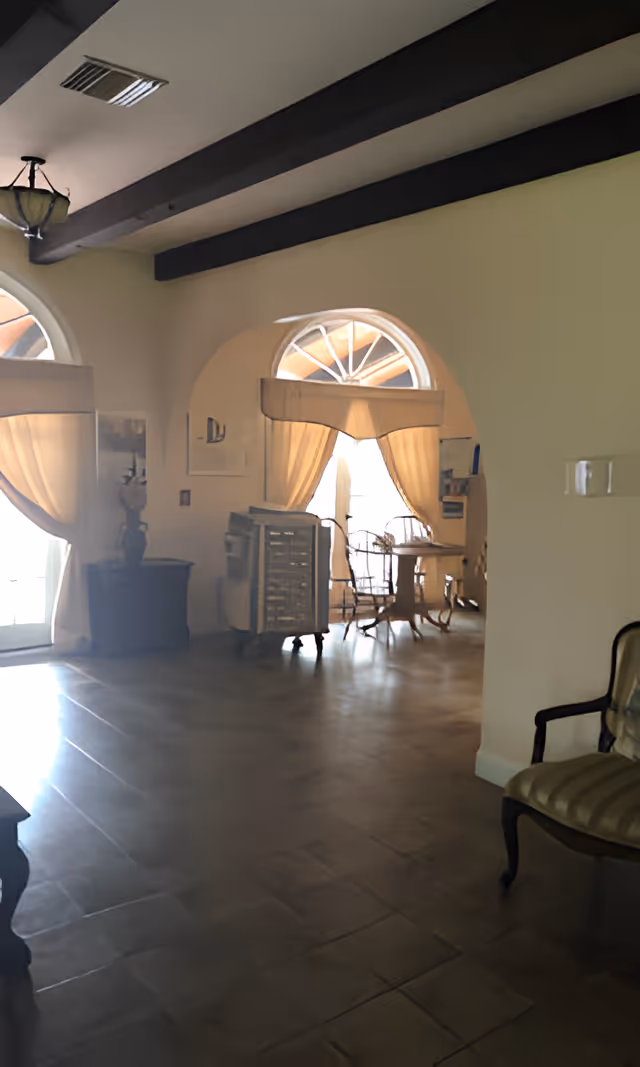 Interior view of a room with arched windows covered by curtains, wooden beams on the ceiling, a small table with chairs near the window, and a cushioned armchair on the right side.