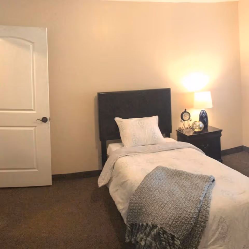 A small bedroom with a single bed featuring a dark headboard, white bedding, and a gray throw blanket. Next to the bed is a dark wooden nightstand with a table lamp, a decorative clock, and a small decorative item. The room has beige walls, a closed white door, and brown carpeted floor.