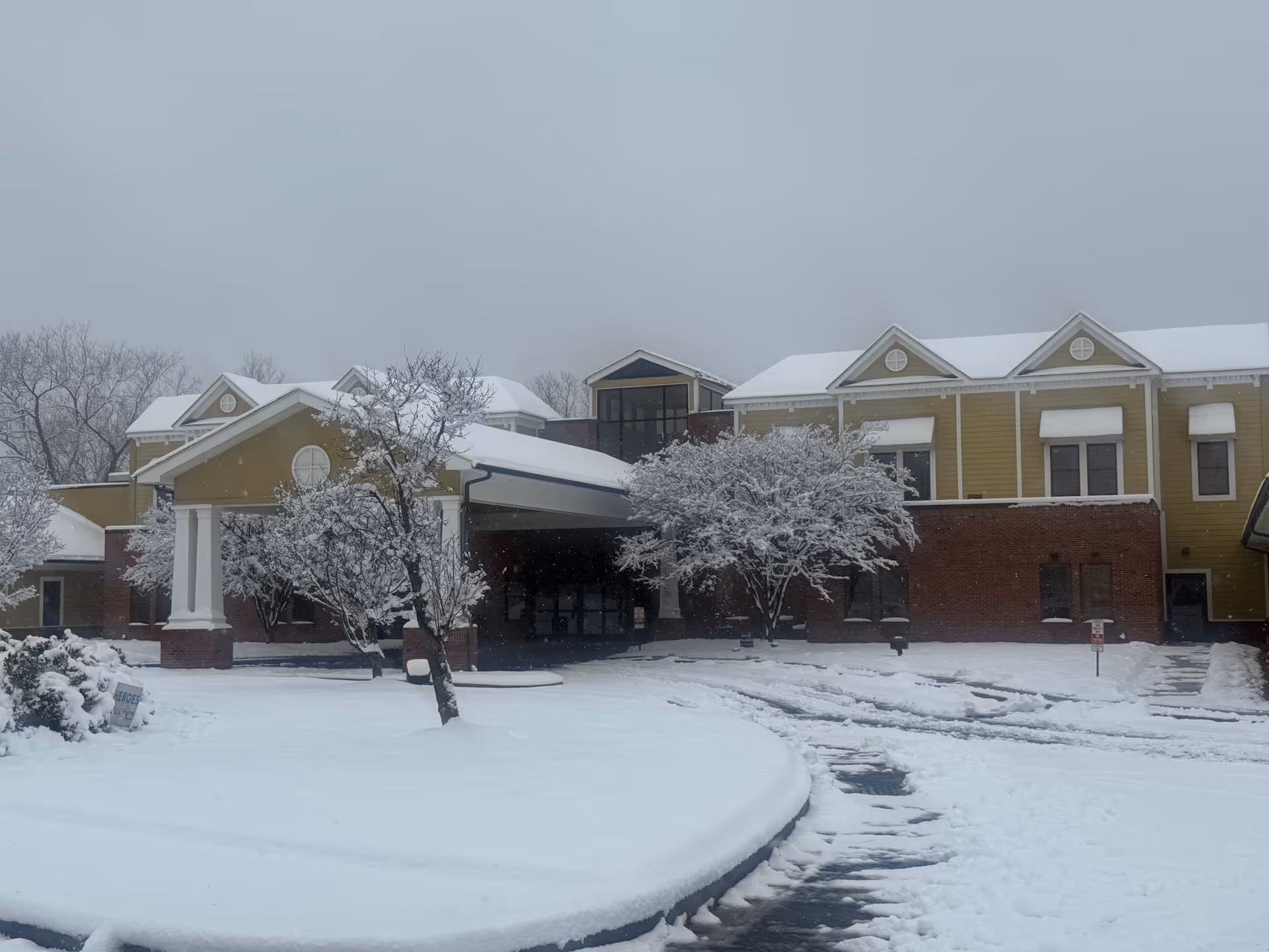 Exterior view of Harborview Post Acute facility covered in snow. The building has a combination of brick and yellow siding with white trim and multiple gabled roofs. Snow-covered trees and a snow-covered driveway are visible in front of the building under a gray, overcast sky.