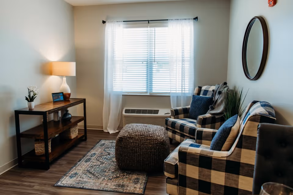 Cozy sitting area with two plaid armchairs, a woven ottoman, a wooden console table with lamp and decor, and a window with sheer curtains.