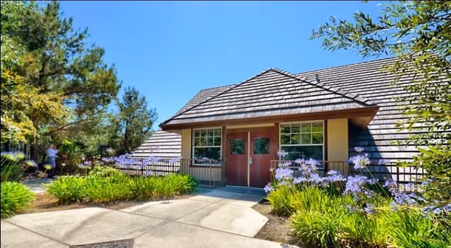 Low-slung building entrance with double doors surrounded by landscaped flower beds, trees, and a clear blue sky.