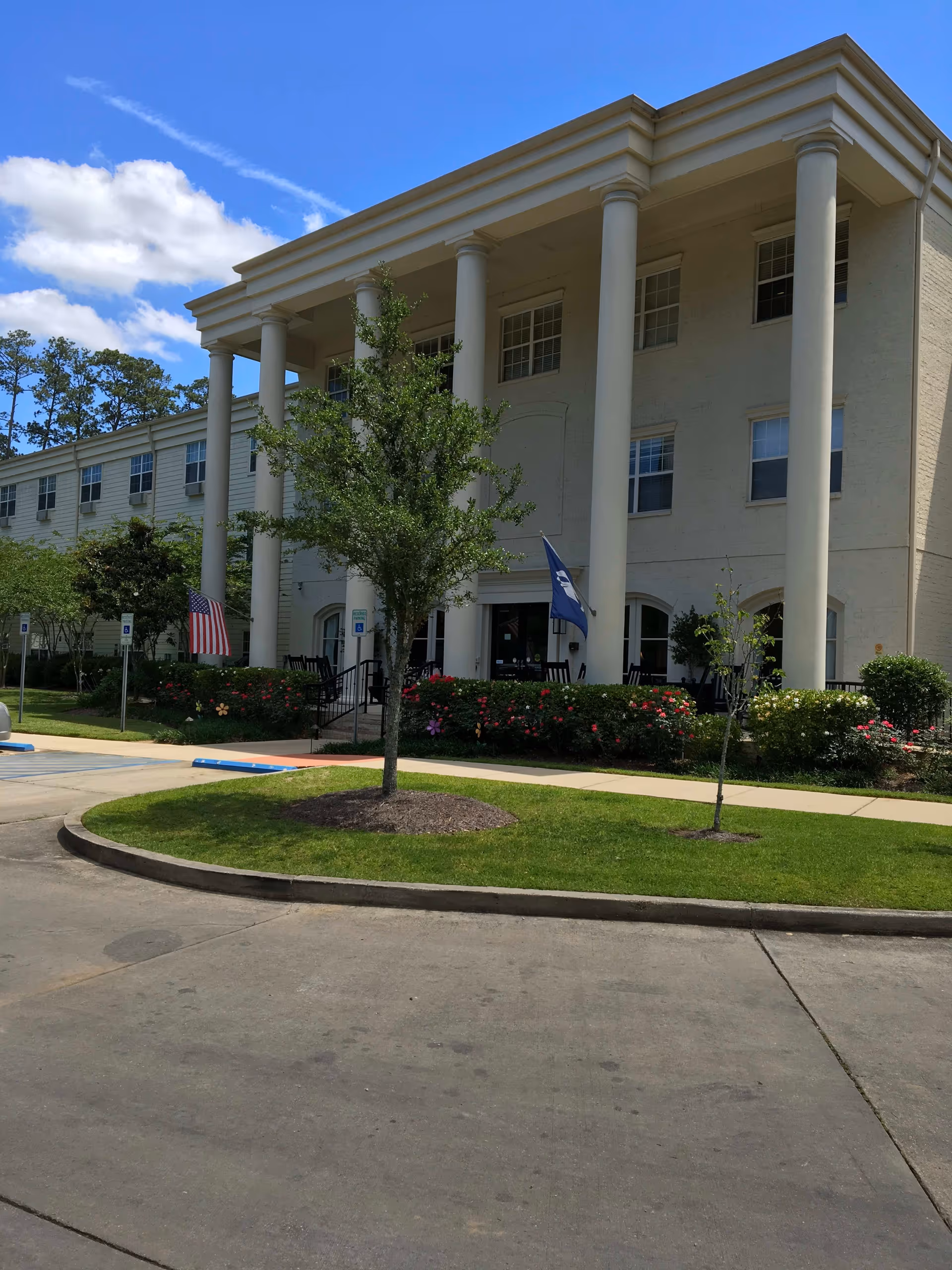 Exterior view of a senior living facility named The Trace, featuring a large building with tall white columns at the entrance, manicured green lawn, small trees, and flower beds. Two flags are displayed near the entrance, and the sky is partly cloudy with blue patches.