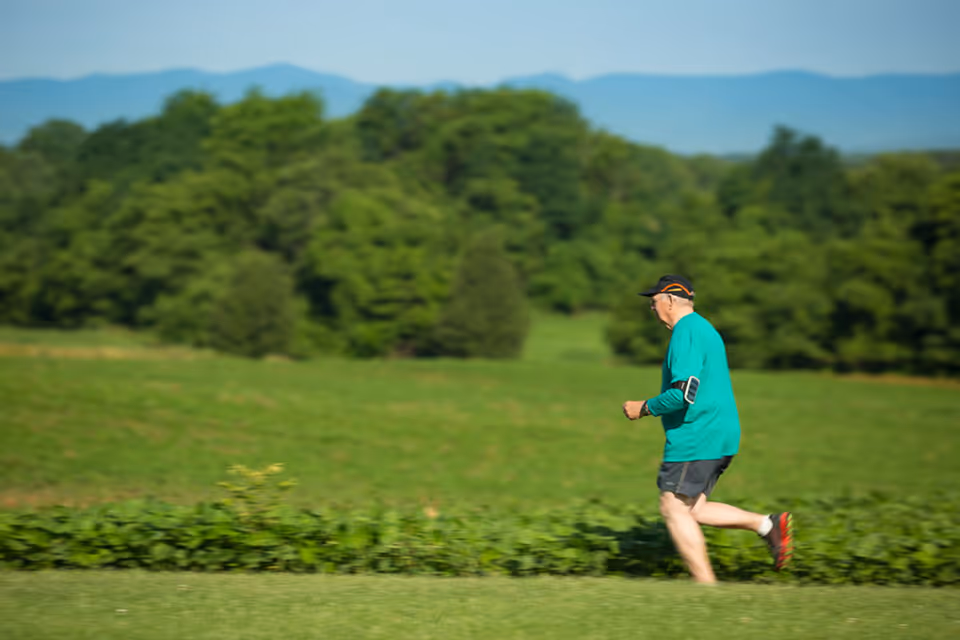 An elderly man wearing a teal shirt, black shorts, and a black cap is jogging outdoors on a grassy field with trees and mountains in the background.