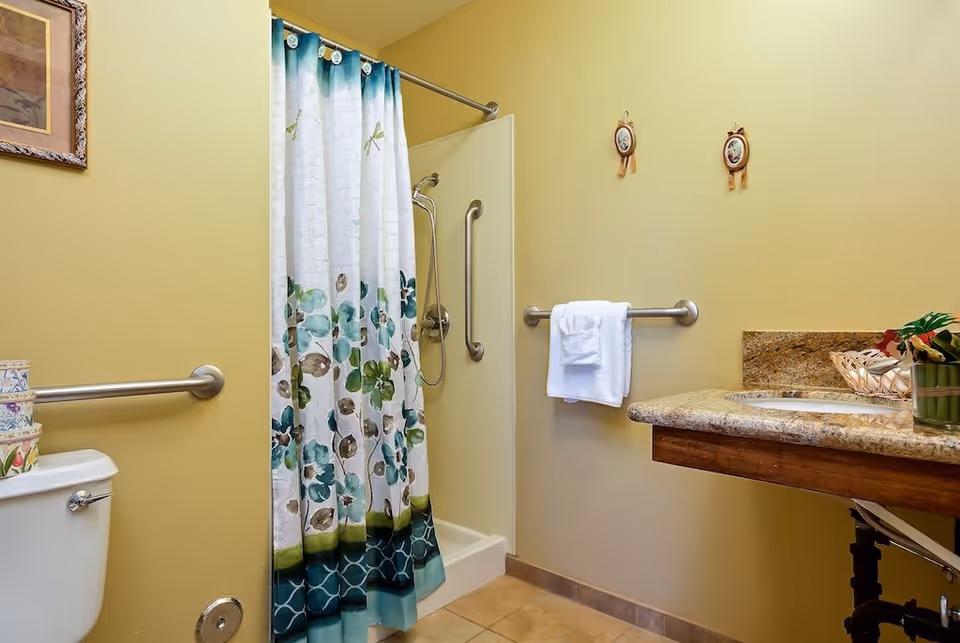 A bathroom with a shower area featuring a floral shower curtain, grab bars on the walls, a white toilet with a grab bar beside it, a granite countertop with a sink, and a towel hanging on a grab bar. The walls are painted yellow and there are two small decorative wall hangings above the towel.